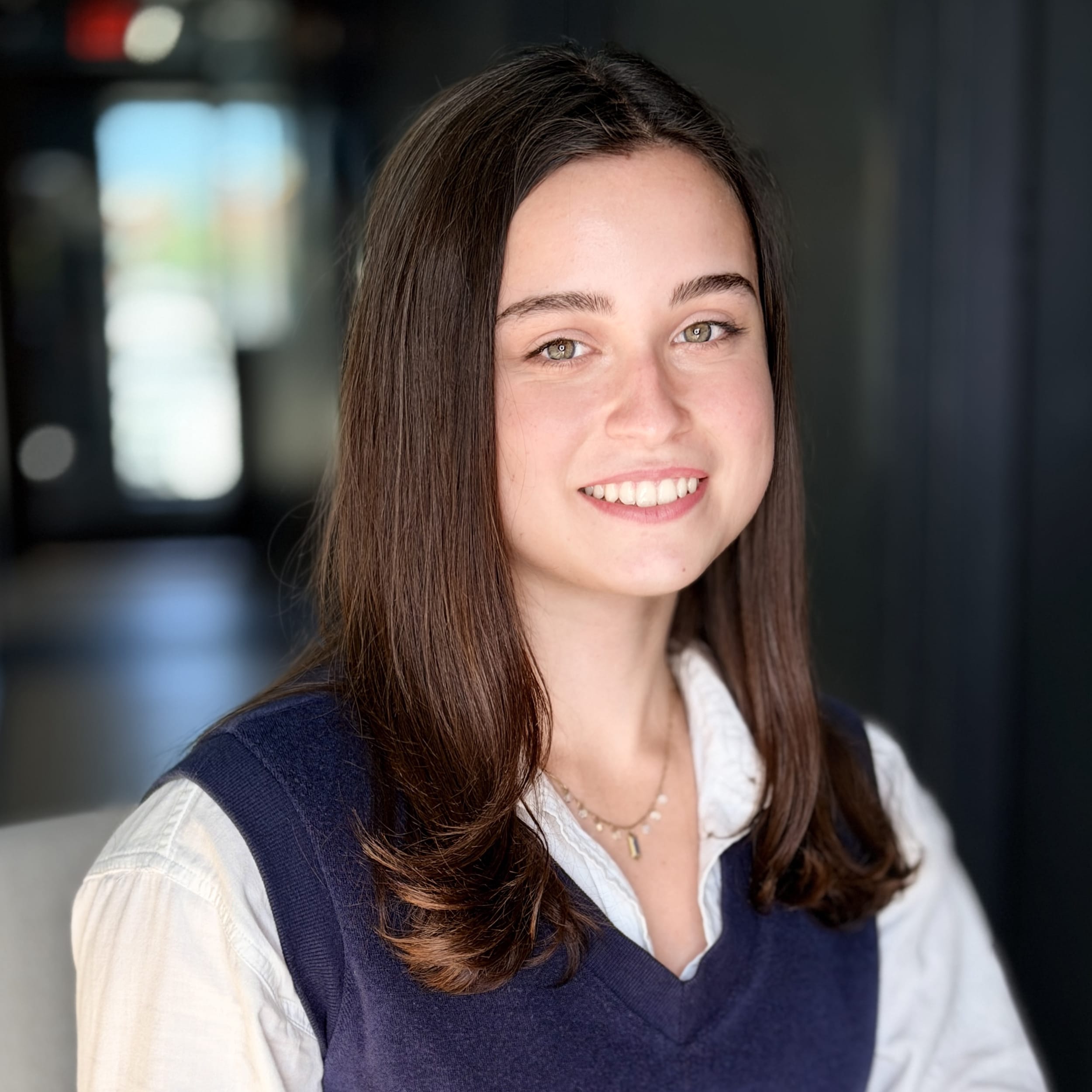Close up of a young, smiling woman with brown hair wearing a navy vest and white collared shirt.